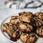 A plate of samoas cookies dipped in chocolate and sprinkled with toasted coconuts. There is a bit on top.