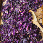 Roasted purple cabbage is fully cooked on a cookie sheet, and topped with sesame seeds with a wooden spoon scooping some up.