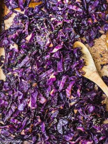 Roasted purple cabbage is fully cooked on a cookie sheet, and topped with sesame seeds with a wooden spoon scooping some up.