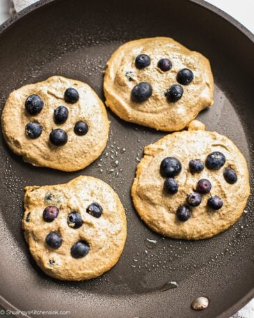 Vegan oatmeal pancakes are heating on a frying pan. There are some extra fresh blueberries on top.