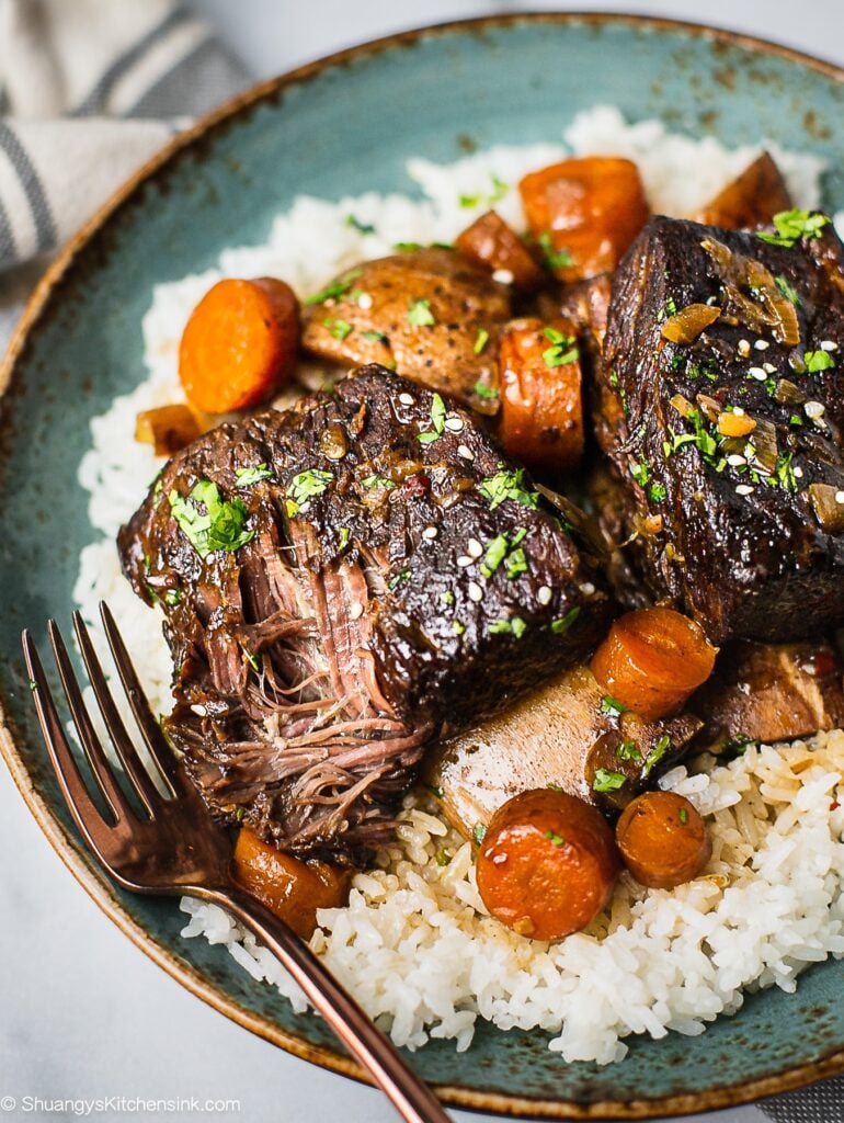 En tallerken med slow cooker koreanske short ribs serveret med jasminris. Der er gulerødder, kartofler i og garneret med koriander... Der er en gaffel, der får et stykke oksekød, der er ved at falde af benet.