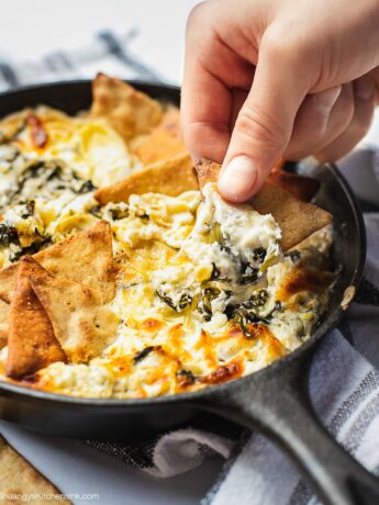 A cast iron skillet full of baked spinach artichoke dip. There is a hand holding a pita chip dipping in.