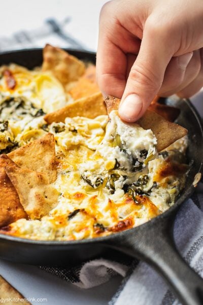 A cast iron skillet full of baked spinach artichoke dip. There is a hand holding a pita chip dipping in.