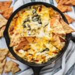 A cast iron skillet full of healthy spinach artichoke dip. There is a hand holding a pita chip dipping in.