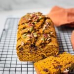 A loaf of healthy pumpkin bread sliced on a cooling rack. There are pumpkin seeds and pecans on top.