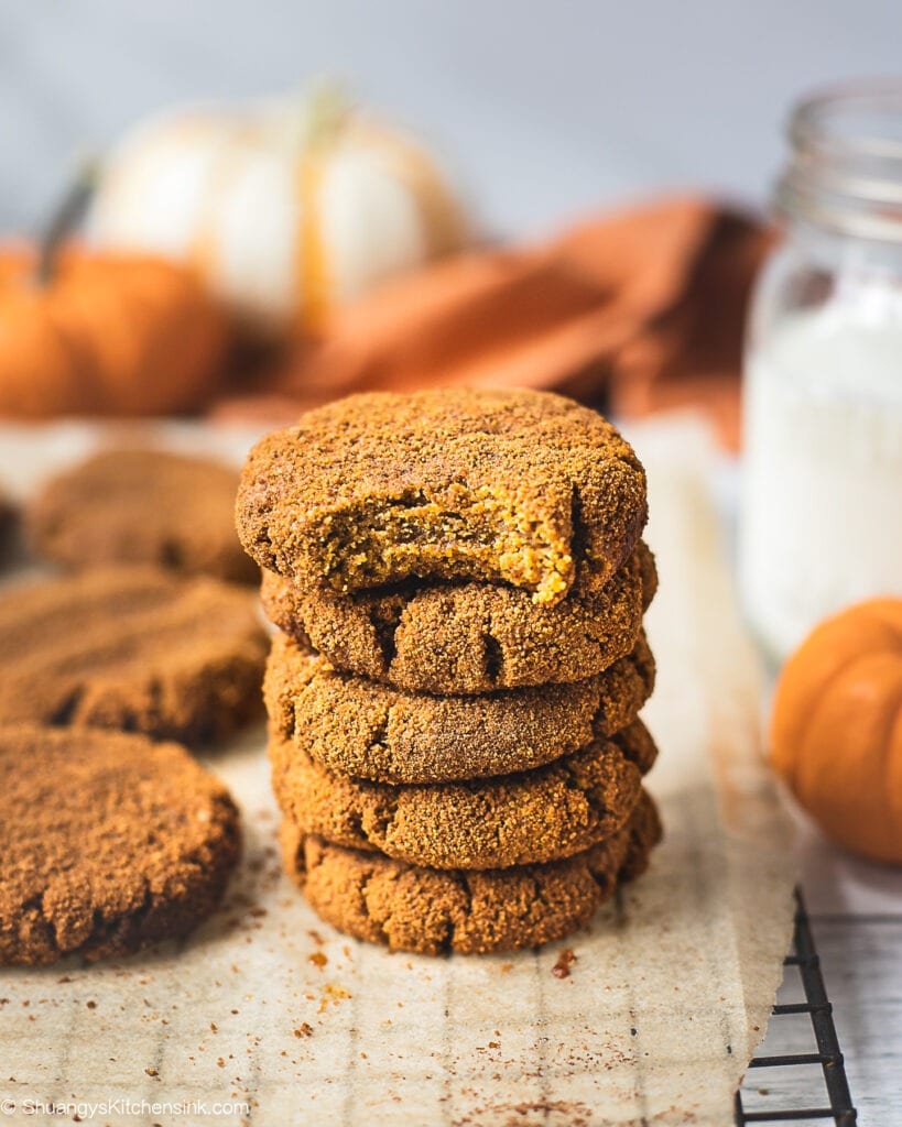 A stack of healthy pumpkin cookies on a piece of parchment paper. There is a glass of milk 2 little pumpkins in the background. The pumpkin spice cookie on top has a bite in it which make it look like a soft pumpkin cookie.