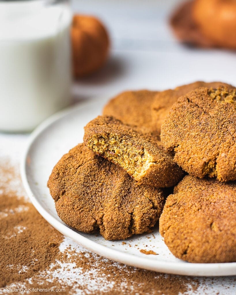 A plate of pumpkin snickerdoodle cookies coated with cinnamon sugar. There is a glass of milk and 2 little pumpkins in the background. There is also a bite into one of the pumpkin cookies that appear to be soft and chewy in texture.