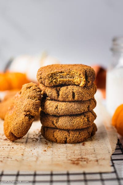 A stack of healthy pumpkin cookies on a piece of parchment paper. There is a glass of milk 2 little pumpkins in the background. The pumpkin spice cookie on top has a bite in it which make it look like a soft pumpkin cookie.