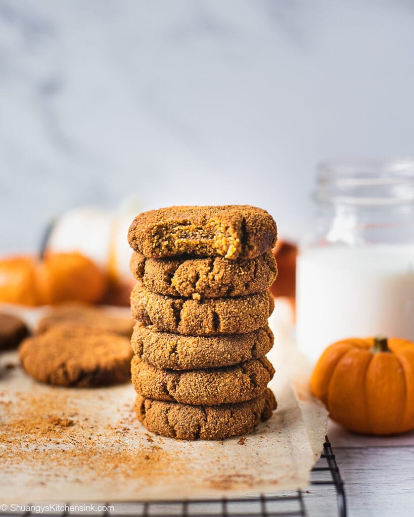 A stack of healthy pumpkin cookies on a piece of parchment paper. There is a glass of milk 2 little pumpkins in the background. The pumpkin spice cookie on top has a bite in it which make it look like a soft pumpkin cookie.