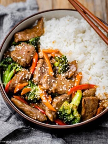 A bowl of beef and broccoli stir fry with broccoli, bell pepper, covered in a glossy savory sauce. Topped with sesame seeds and served with white jasmine rice. There is a pair of chopsticks on the side.