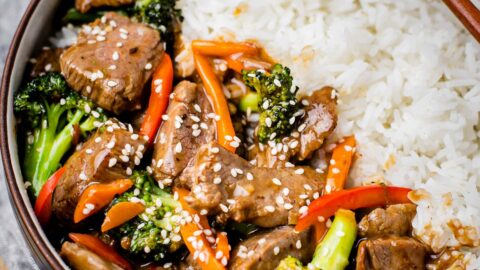 A bowl of beef and broccoli stir fry with broccoli, bell pepper, covered in a glossy savory sauce. Topped with sesame seeds and served with white jasmine rice. There is a pair of chopsticks on the side.