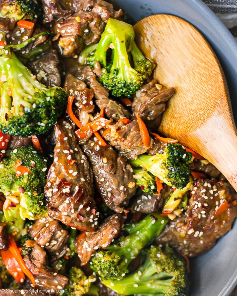 A pan of beef and broccoli stir fry with carrots and sesame seeds on top. There is a wooden spoon inserted in the tender beef flank steak.