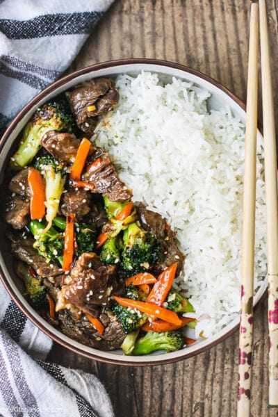 A bowl of healthy beef and broccoli stir fry with rice. there are green onion garnish on top and a pair of chopsticks on the side.