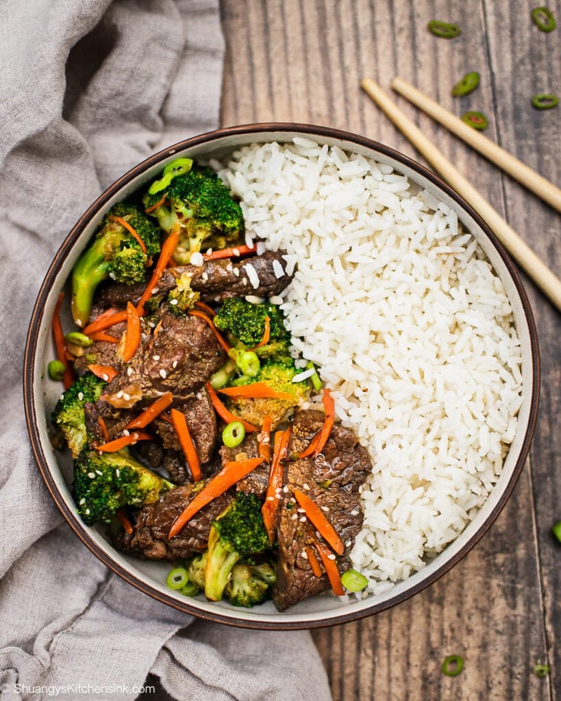 A bowl of healthy beef and broccoli stir fry with rice. there are green onion garnish on top and a pair of chopsticks on the side.