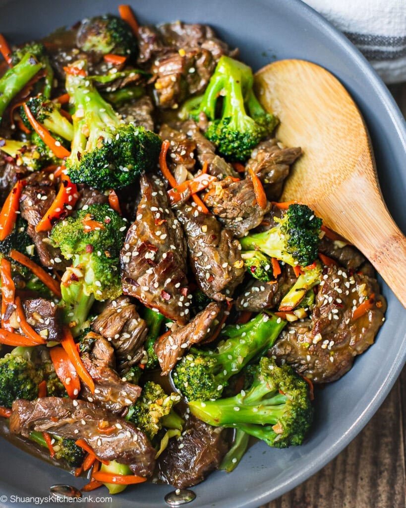 A pan of beef and broccoli stir fry with carrots and sesame seeds on top. There is a wooden spoon inserted in the tender beef flank steak.A pan of beef and broccoli stir fry with carrots and sesame seeds on top. There is a wooden spoon inserted in the tender beef flank steak.