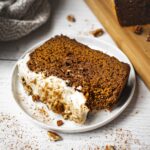 A slice of copycat starbucks gingerbread loaf with healthy cream cheese frosting. There are pecan pieces and cinnamon on top. There is a bite on the gingerbread loaf.