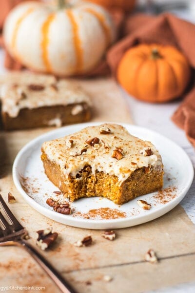 A piece of pumpkin bar with cream cheese frosting on a little plate. There are pumpkin spice and pecans sprinkled on top. There is a bite in the middle. The pumpkin bar appears to be soft and chewy.