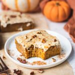 A piece of pumpkin bar with cream cheese frosting on a little plate. There are pumpkin spice and pecans sprinkled on top. There is a bite in the middle. The pumpkin bar appears to be soft and chewy.