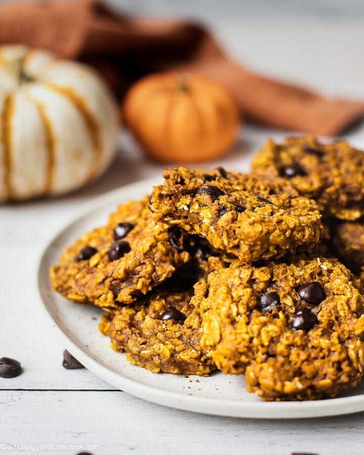 A plate of pumpkin oatmeal cookies with chocolate chips. There are a couple pumpkins in the background.