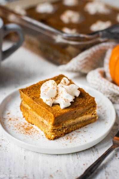A piece of pumpkin pie bars with almond flour crust is on a plate served with dairy free whipped cream. There is a whole plan of pumpkin pie bars in the background.