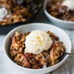 A bowl of caramel apple crisp with soft baked apples, crunchy oat crisp toppings and a scoop of dairy free vanilla ice cream. There is also drizzle of salted caramel sauce on top.