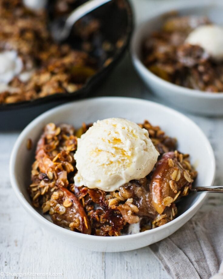 A bowl of caramel apple crisp with soft baked apples, crunchy oat crisp toppings and a scoop of dairy free vanilla ice cream. There is also drizzle of salted caramel sauce on top.