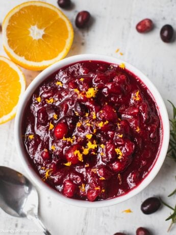 A bowl of healthy cranberry sauce that is topped with orange zest. There are some fresh oranges and cranberries in the background.
