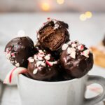 A batch of peppermint chocolate truffles with candy cane are in a mug. The top chocolate energy bite has a bite in it. The texture appears to be gooey and fudgy in the middle.
