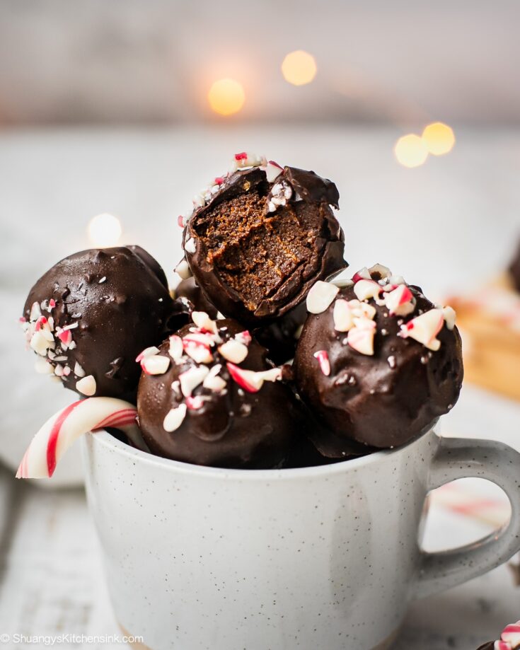 A batch of peppermint chocolate truffles with candy cane are in a mug. The top chocolate energy bite has a bite in it. The texture appears to be gooey and fudgy in the middle.