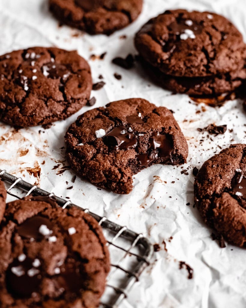 A batch of freshly baked vegan double chocolate cookies. It looks soft and brownie-like with melted dark chocolate chips on top and a sprinkle of sea salt.