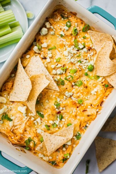 A baking dish full of healthy low carb buffalo chicken dip that is cheesy and creamy. It is being serve by fresh celery sticks and low carb tortilla chips (made with almond flour). There are scallions and blue cheese on top.