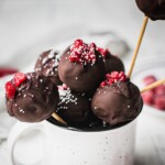 A mug full of raspberry vegan cheesecake balls coated with dark chocolate, and topped with raspberry and sprinkles. There are more vegan cake pops in the back.