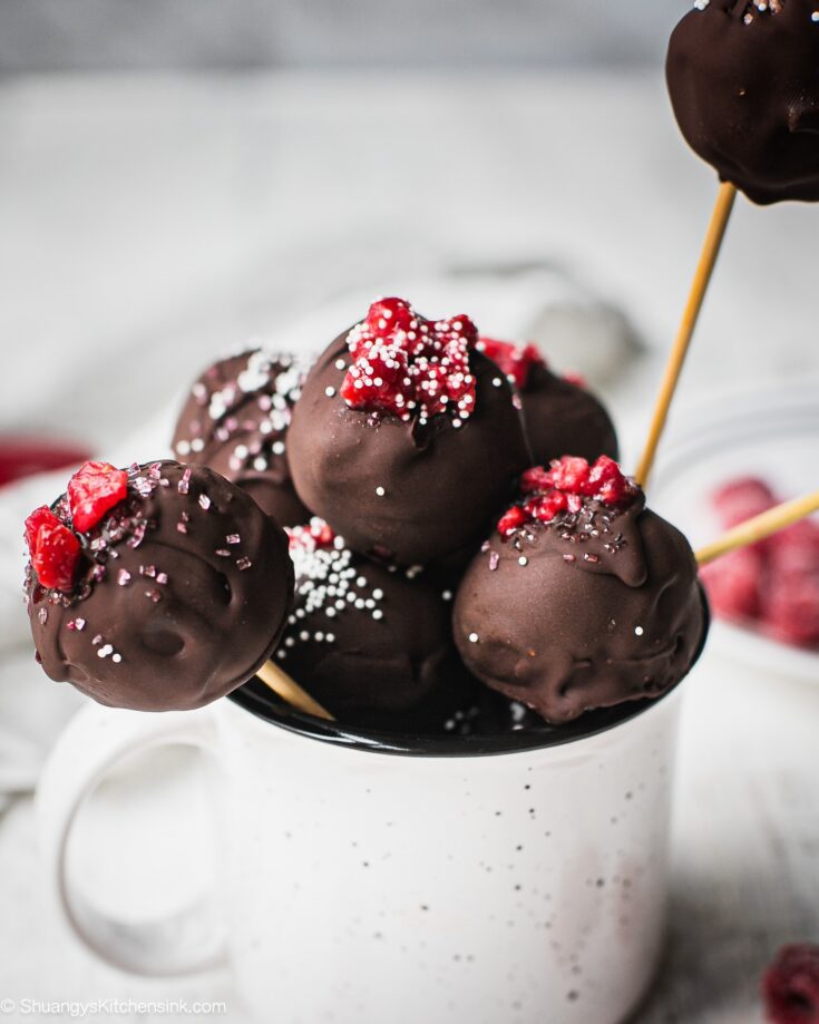 A mug full of raspberry vegan cheesecake balls coated with dark chocolate, and topped with raspberry and sprinkles. There are more vegan cake pops in the back.