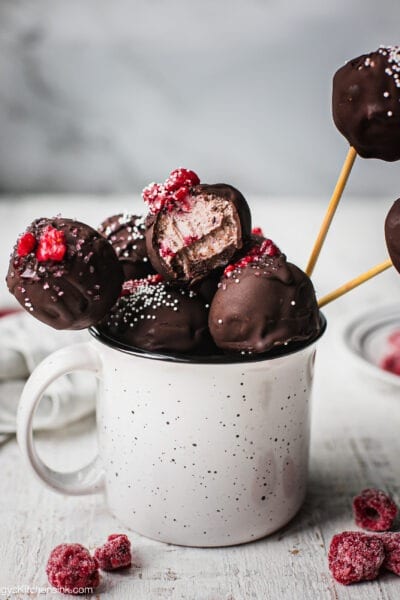 A mug full of raspberry vegan cheesecake balls coated with dark chocolate, and topped with raspberry and sprinkles. There is a bite in one of the chocolate raspberry truffles which shows the texture inside to be smooth and gooey. There are more vegan cake pops in the back.