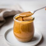 A jar of vegan caramel sauce with a spoonful of it coming out of the jar. The texture appears to be thick and velvety and the color is golden brown.