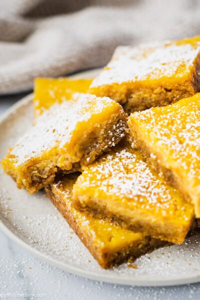 A plate of gluten free lemon bar that has powdered sugar on top and a bite in the middle. The texture appears to be creamy and soft.