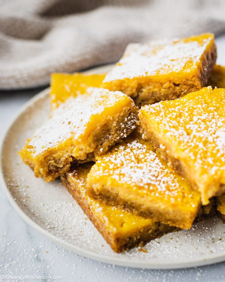 A plate of gluten free lemon bar that has powdered sugar on top and a bite in the middle. The texture appears to be creamy and soft.