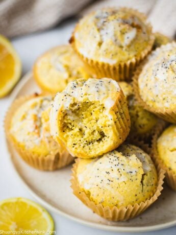 A batch of healthy lemon poppy seed muffins on a plate. There is lemon coconut icing on top of these healthy muffins. There is a bite in the muffins and the texture appears to be fluffy and moist.