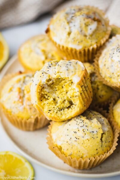 A batch of healthy lemon poppy seed muffins on a plate. There is lemon coconut icing on top of these healthy muffins. There is a bite in the muffins and the texture appears to be fluffy and moist.