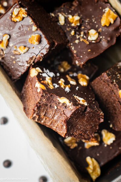 A batch of no bake brownies in a parchment paper lined baking dish. There is a bite in the middle. The texture appears to be fudgy and chewy