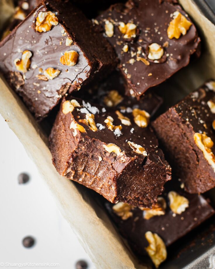 A batch of no bake brownies in a parchment paper lined baking dish. There is a bite in the middle. The texture appears to be fudgy and chewy