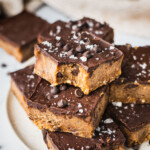 There is a plate full of gluten-free cheesecake bars. The healthy dessert is topped with a creamy dairy-free chocolate ganache, chocolate chips and a sprinkle of sea salt.