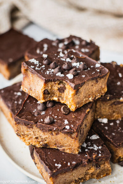 There is a plate full of gluten-free cheesecake bars. The healthy dessert is topped with a creamy dairy-free chocolate ganache, chocolate chips and a sprinkle of sea salt.