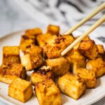 A plate full of air fried crispy tofu. One piece is being picked up by chopsticks