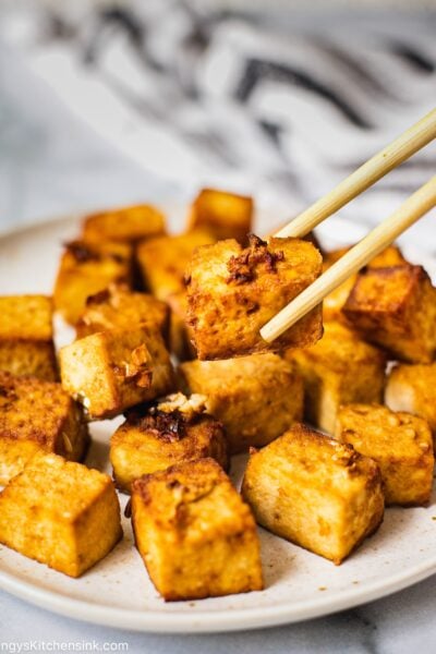A plate full of air fried crispy tofu. One piece is being picked up by chopsticks