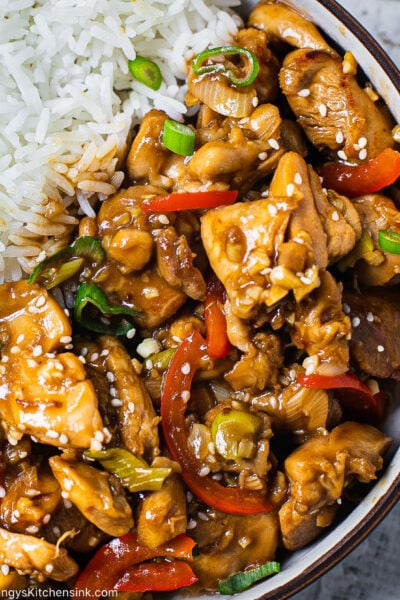 A bowl of jasmine rice and Healthy Chinese Garlic Chicken. There are red peppers and green scallions as well and a sprinkle of sesame seeds