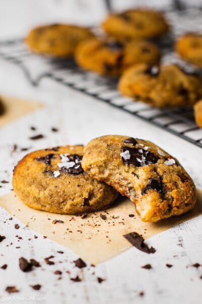 two cookies on a counter top. Someone has taken a bite from one of the gluten free cookies. These are vegan chocolate and sea salt on the cookies. In the background there are more cookies on a cooling wrack.