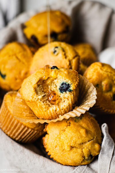 Honey is being drizzled on a gluten free cornbread muffin. You can see that there are fresh blueberries in the baked good as well.