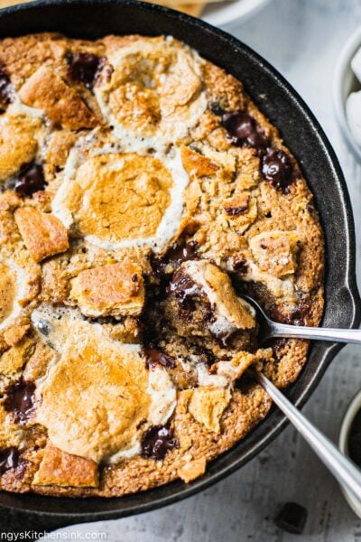 two spoons have been left inside a warm gluten free S’mores Stuffed Cookie Skillet that is topped with chocolate and caramelized marshmallows