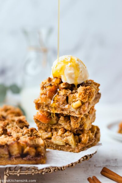 paleo apple pie crumble bars stacked on top of each other with a dollop of vegan ice cream and maple syrup is being drizzles on top.